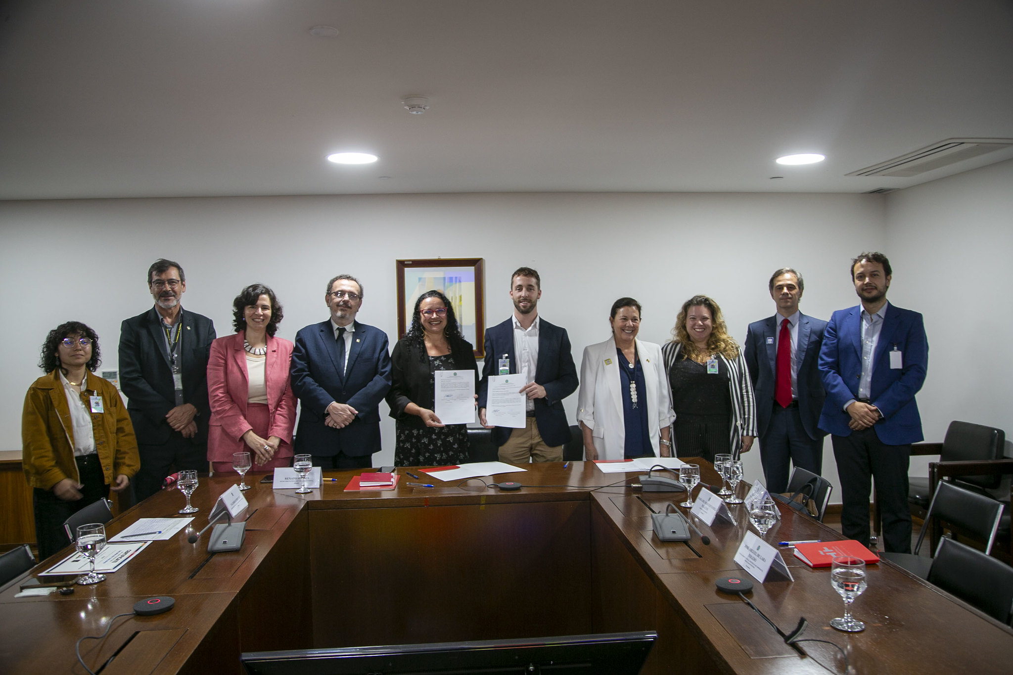 A group of ten people stand smiling behind a wooden conference table in a meeting room, with two people in the center holding up signed documents.
