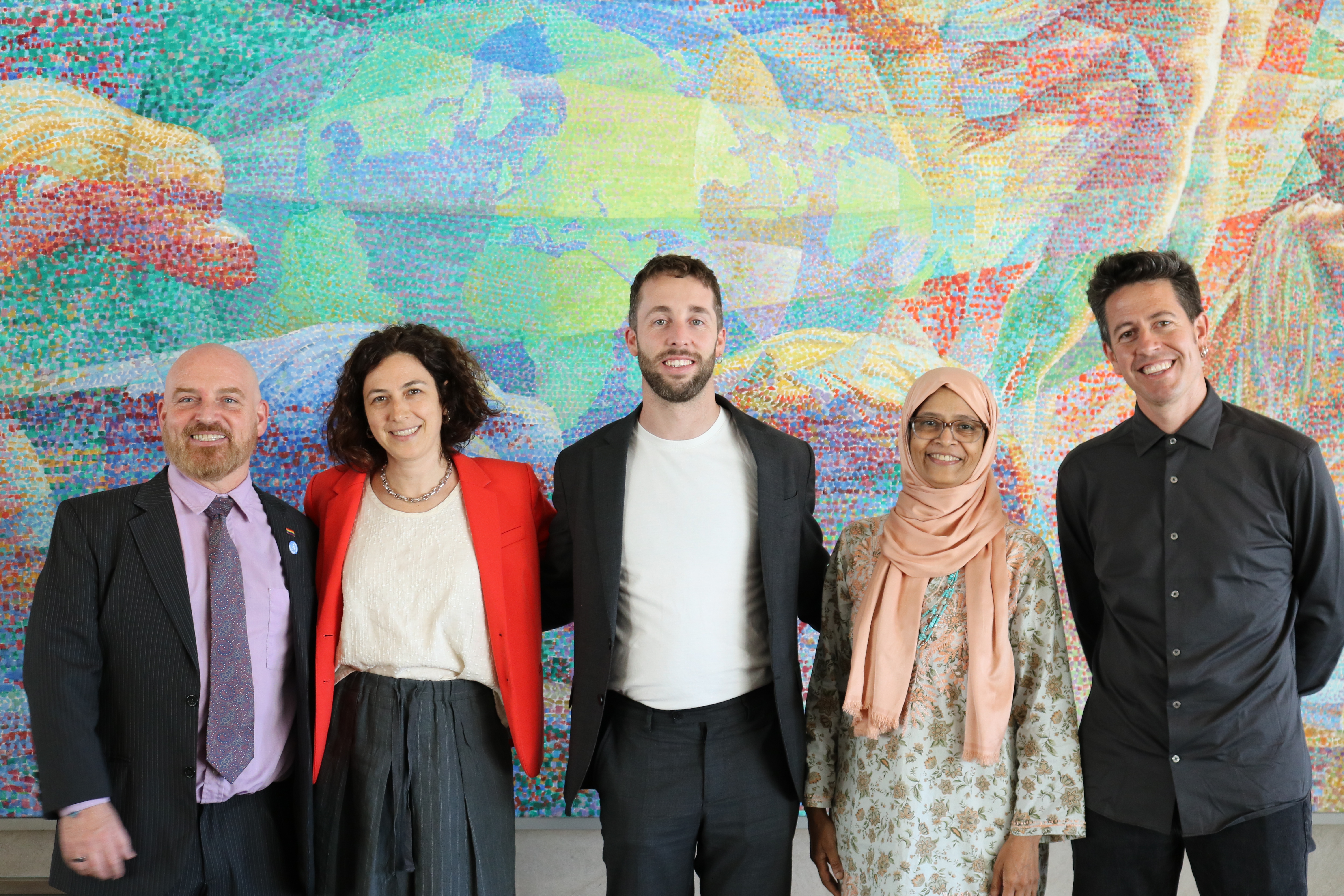 Five people stand smiling in front of a large, colorful mosaic mural depicting a globe and abstract figures.