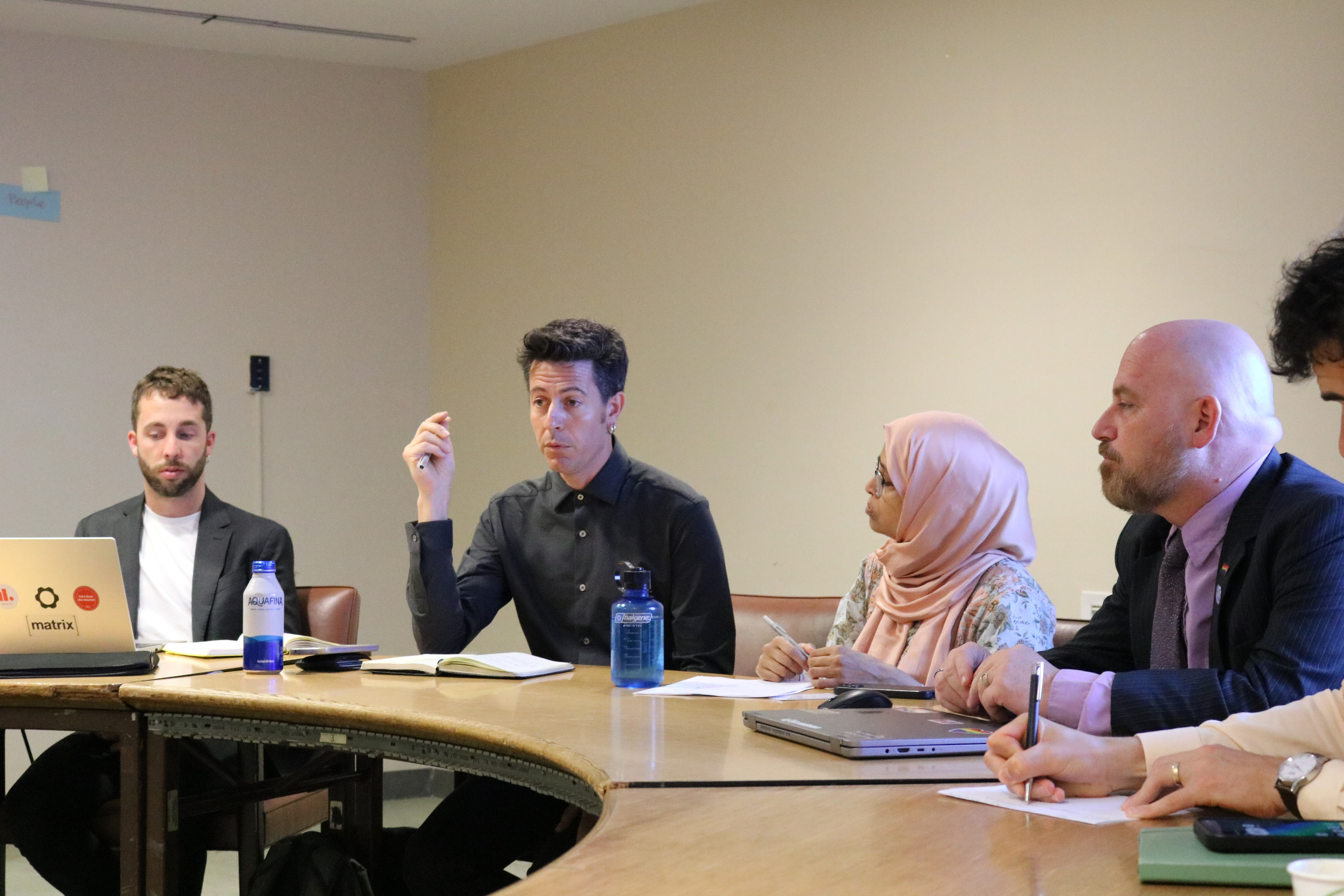 A man gestures while speaking during a meeting at a wooden table, with several other people seated beside him listening and taking notes.